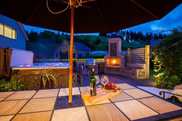 Evening view of an outdoor entertaining terrace beneath a large umbrella, with a tiled dining table in the foreground, wine and food, a built-in outdoor fireplace, trellis screen, garden planting, and a spa or hot tub visible to the left. Warm firelight and blue twilight create an inviting ambience.