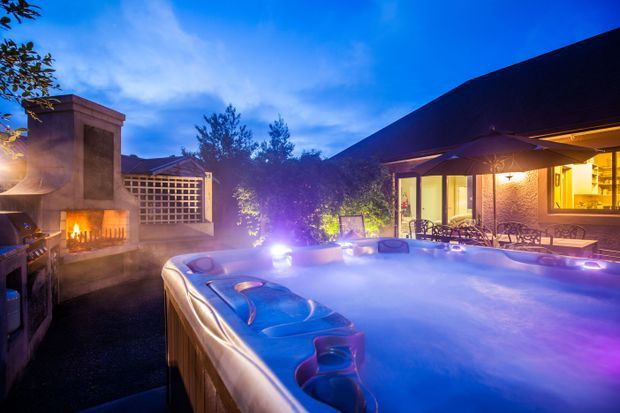 Twilight view of a residential outdoor entertaining area dominated by a large illuminated spa pool, with an outdoor fireplace, paved terrace, landscaped planting, and a warmly lit single-storey house with a pitched roof in the background.