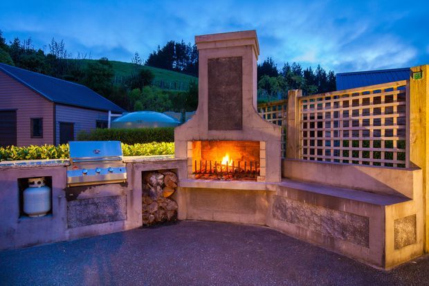An outdoor entertaining area featuring a built-in masonry fireplace with a lit fire, integrated seating, a stainless-steel barbecue and gas storage, timber lattice screening, and stacked firewood. The setting appears to be a landscaped residential garden at dusk, with a house and sloping greenery in the background.