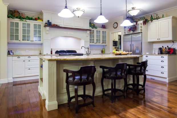 A spacious traditional kitchen with extensive white raised-panel cabinetry, glass-front display cupboards, a large central island with a pale stone benchtop and seating for four, and polished dark timber flooring. Decorative plants, hanging pendant lights, dark timber bar stools, and classic detailing create a warm, refined atmosphere.