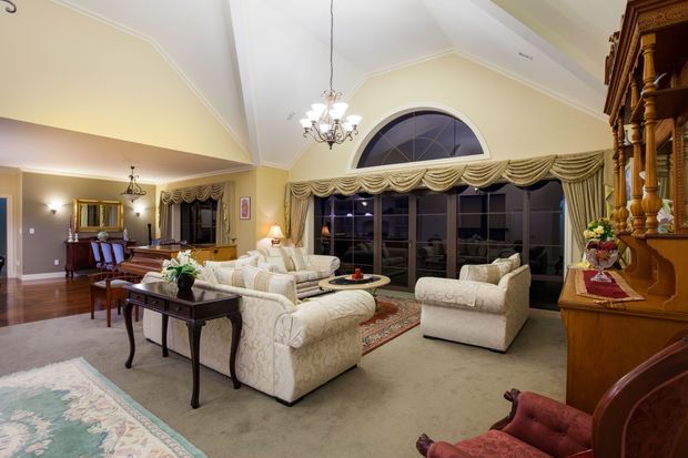A spacious, traditionally styled living room with cream upholstered sofas and armchairs arranged around a round coffee table, set over wall-to-wall carpet and layered area rugs. The room features polished timber flooring in the adjoining dining area, ornate draped floor-to-ceiling windows with a large arched transom, decorative chandeliers and wall sconces, classical crown moulding, and substantial dark timber built-in cabinetry or shelving along the right side. A grand piano and dining furnishings are visible in the open-plan adjoining area.