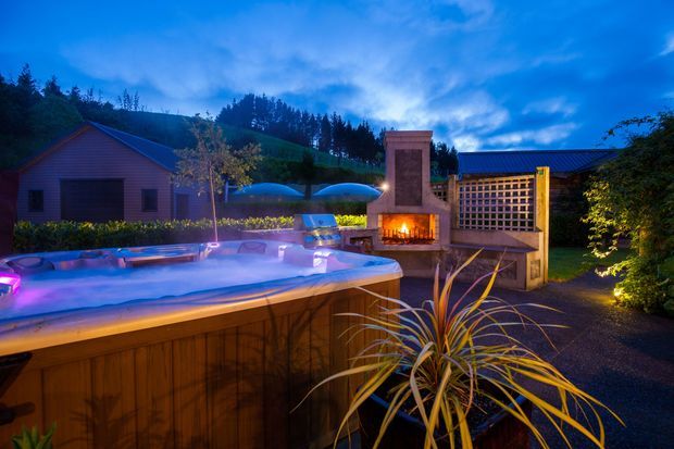 Twilight exterior view of a landscaped backyard entertainment area featuring a raised timber-clad spa or plunge pool in the foreground, an outdoor fireplace with a timber pergola or privacy screen, and a small garden structure with a pitched roof in the background. Warm firelight and landscape lighting contrast with the blue evening sky.