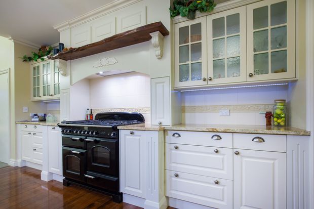 Traditional-style kitchen with extensive white raised-panel cabinetry, a large black freestanding range, glass-front upper cabinets, a decorative tiled backsplash, and a substantial dark timber display shelf above the cooking area. Polished timber flooring and warm lighting create a classic, refined atmosphere.