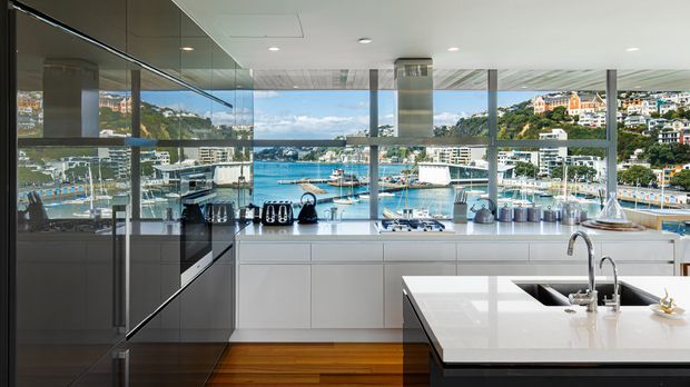 Contemporary kitchen with sleek white handleless cabinetry, a large white island with integrated sink and tapware, dark reflective cabinetry, timber flooring, and expansive floor-to-ceiling glazing overlooking a marina and waterfront landscape. The space is bright, polished, and highly connected to the view.