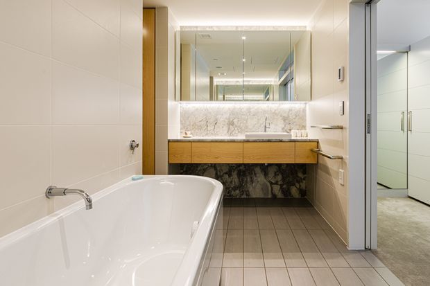 A bright contemporary bathroom featuring a freestanding white bathtub, large-format light wall tiles, grey tiled flooring, a floating timber vanity with a white-and-grey marble backsplash and dark stone-look lower wall, and a wide mirrored cabinet. An open doorway reveals a walk-in wardrobe or adjacent storage area.