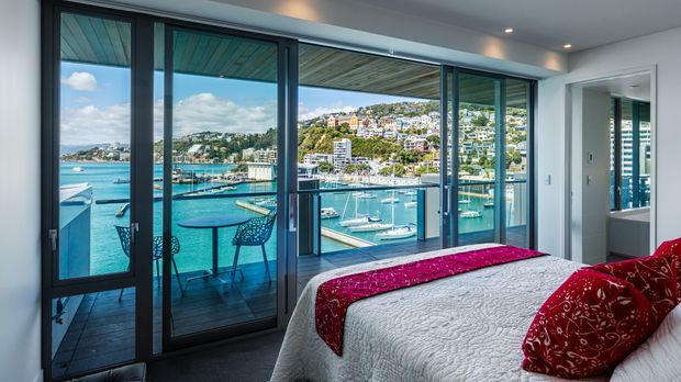 A bright modern bedroom overlooking a marina and turquoise waterfront through expansive floor-to-ceiling sliding glass doors. The room has a white bed with a textured cover, vivid red patterned cushions and runner, dark-framed glazing, and a small balcony with café seating. A bathroom is partially visible through the opening at right.