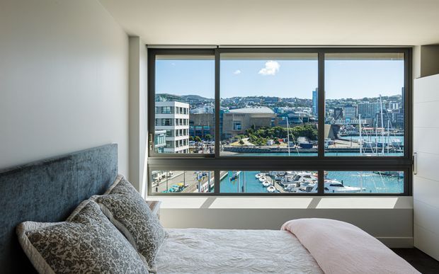 Bright contemporary bedroom with a large floor-to-ceiling framed window overlooking a marina, waterfront buildings and a hilly urban landscape. The room features a built-in upholstered grey headboard, white bedding, patterned cushions, a pale pink throw, white walls and built-in white cabinetry.