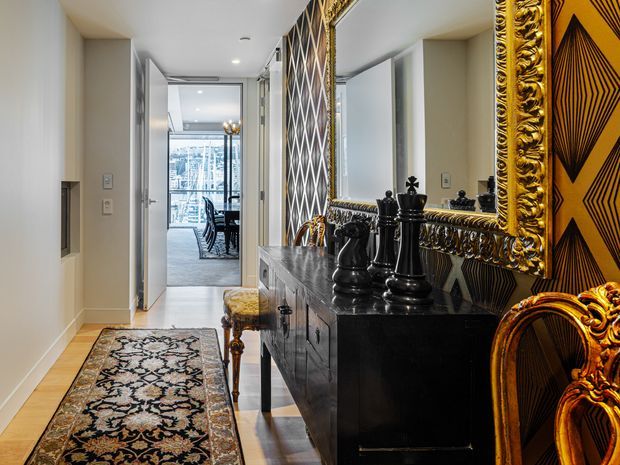 A refined hallway or entry passage featuring a black console cabinet topped with ornate black decorative objects, an oversized gilded mirror, and richly patterned black-and-gold wall panels. A traditional patterned runner extends along the light timber floor, while an open doorway leads toward a dining or living area with extensive glazing. Ornate gold-framed seating is partially visible in the foreground.