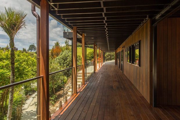 A long covered exterior walkway or veranda with dark timber decking, timber posts and wall cladding, black metal balustrades, and exposed roof framing. Glazed windows line the right side, while lush tropical landscaping and a sloping garden are visible beyond the open railing.