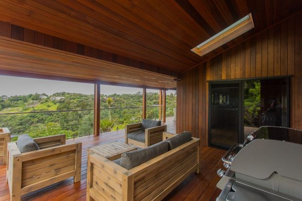 A covered timber-clad outdoor living area or balcony overlooking a lush landscape. The space features warm wood wall and ceiling lining, timber decking, large glazed openings with dark frames, built-in or substantial timber lounge seating, a coffee table, and a barbecue at the right. A roof skylight brings additional daylight into the sheltered entertaining area.