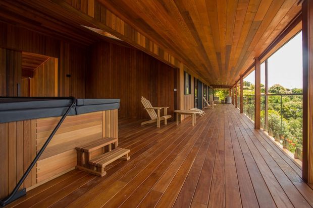 A long, covered timber deck or veranda with extensive timber wall, ceiling, and flooring, creating a warm outdoor entertaining space. A built-in bar or counter occupies the left side, with simple timber lounge chairs and a small table along the deck. Open glazing and railing on the right overlook dense greenery and a sunny landscape.