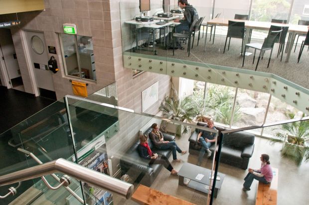 Multi-level contemporary commercial interior viewed from a stairway, showing a glazed balustrade, lounge seating in a lower reception area, and an upper open dining or breakout space with tables and chairs. Exposed light brick walls, dark flooring, extensive glazing, and visible structural metalwork define the space.