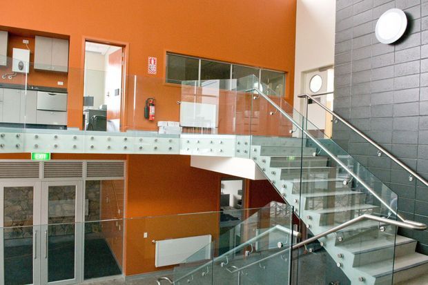 Interior view of a contemporary multi-level commercial or institutional lobby with a prominent glass-and-metal staircase, glass balustrades, and a mezzanine walkway. The space features vivid orange painted walls, dark grey masonry wall cladding, pale tiled flooring, extensive glazing, and exposed circulation routes. A fire extinguisher, exit doors, and accessible upper-level workspace are visible.