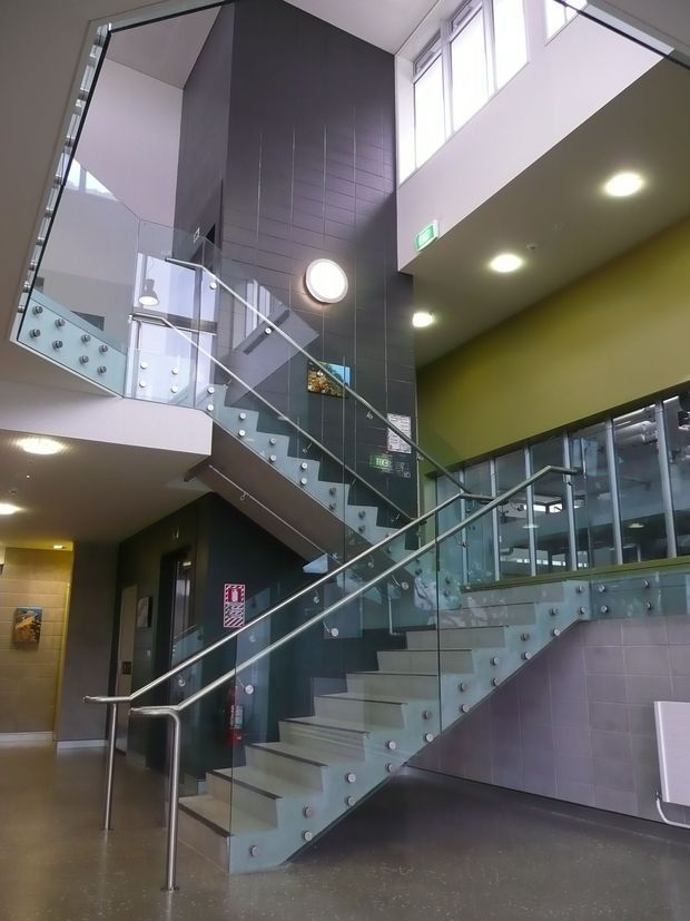 Interior view of a contemporary multi-level public or commercial building lobby, dominated by a sculptural stairway with pale tiled steps, glass balustrades, and stainless-steel handrails. Dark grey tiled wall panels, olive-green painted walls, exposed upper-level glazing, recessed ceiling lights, and visible emergency signage create a bright, functional institutional atmosphere.