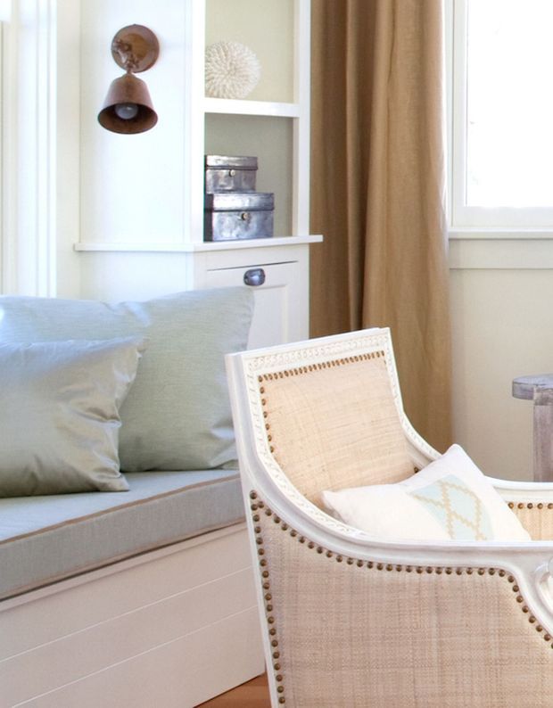 Bright, softly styled bedroom corner featuring a built-in window seat with pale blue cushions, white built-in cabinetry and shelving, a woven cane armchair with nailhead trim, beige curtains, and a small vintage wall sconce. The room has a light, relaxed coastal Hamptons character.