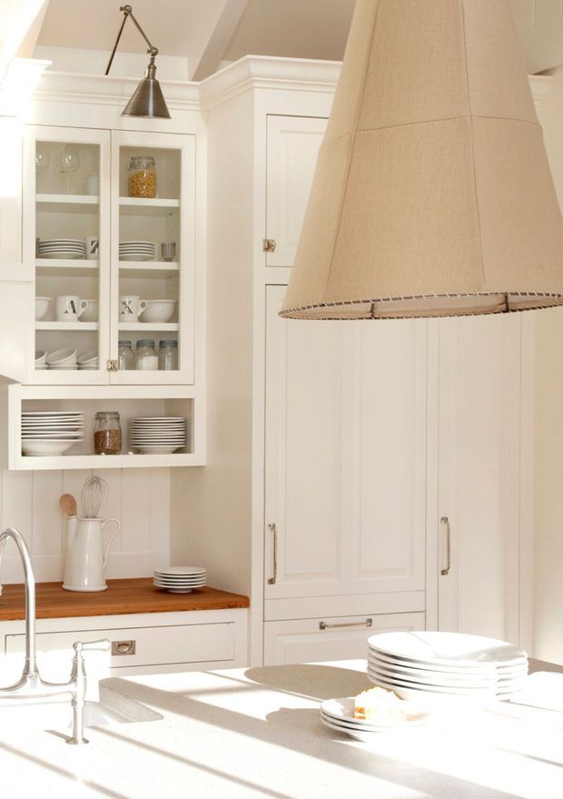 Bright, traditional-style white kitchen with detailed shaker cabinetry, glass-front display cupboards, open shelving, a timber benchtop section, pale stone island, farmhouse-style sink tapware, and an oversized fabric pendant light. Sunlight creates strong highlights across the island and cabinetry.