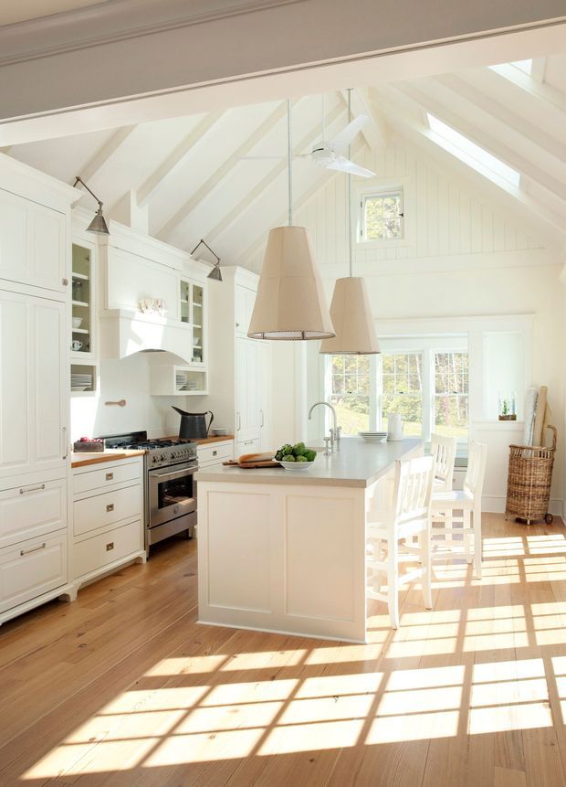 Bright, spacious white kitchen with a large central island, integrated cabinetry, stainless steel range, pendant lights, and white bar stools. The room has a steep vaulted, white-painted timber-panelled ceiling with exposed rafters, skylights, and clerestory-style glazing. Warm timber flooring and abundant sunlight create an airy, inviting atmosphere.
