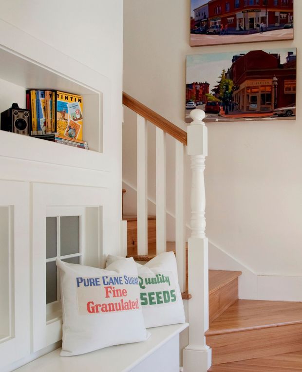 A bright, traditional stairway and landing with white painted built-in wall niches, a small bench seat with printed cushions, a vintage camera and Tintin books, timber stair treads, and a white turned balustrade with timber handrail. Two colourful framed architectural or streetscape artworks hang on the wall.