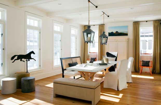 Bright, refined dining room with white-painted walls and trim, tall multi-pane windows, warm timber flooring, and a long dining table surrounded by upholstered seating. Hanging lantern-style pendant lights, draped windows, a fireplace mantel, and an equestrian sculpture contribute to a classic coastal-inspired interior.