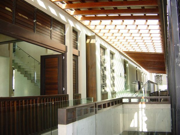 A bright, contemporary interior circulation space with a long covered atrium or hallway, exposed timber roof beams and slatted skylight structure, timber louvres, pale rendered walls, glass balustrades, and a prominent timber entry door. A staircase and multiple levels are visible, creating a strong sense of openness and architectural rhythm.