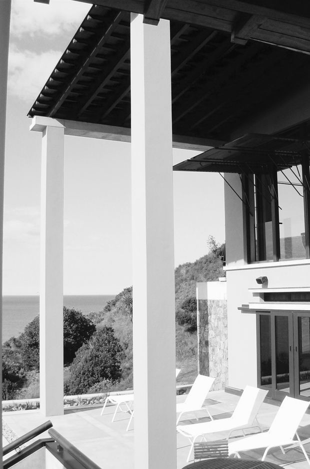 Black-and-white architectural photograph of a modern coastal house terrace. Tall white square columns support a deep exposed-beam roof or pergola, with outdoor sun loungers on a paved terrace. The house has large glazed doors and windows, stone wall cladding, and a hillside setting overlooking the sea.