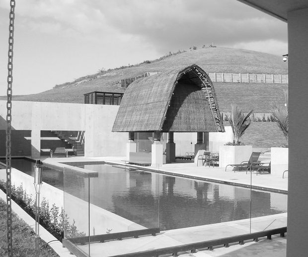 Monochrome architectural photograph of a contemporary outdoor pool courtyard. A long rectangular swimming pool is enclosed by frameless glass balustrades and surrounded by pale concrete paving, lounge chairs, planters, and sculptural palms. The main feature is a curved, thatched or woven canopy over an outdoor seating area, set against a smooth concrete wall and a landscaped hillside. The image is viewed from beneath an overhanging roof or balcony, with a hanging chain visible in the foreground.