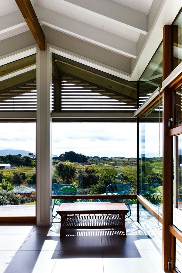 A bright, glazed interior sunroom or enclosed outdoor sitting area with expansive floor-to-ceiling windows overlooking a landscaped coastal or rural setting. The room has a high vaulted ceiling with exposed dark timber beams, white trim and ceiling boards, pale tiled flooring, large glass surfaces, and warm timber-framed doors. A simple timber coffee table and woven outdoor-style chairs occupy the space, with strong sunlight creating striped shadows.