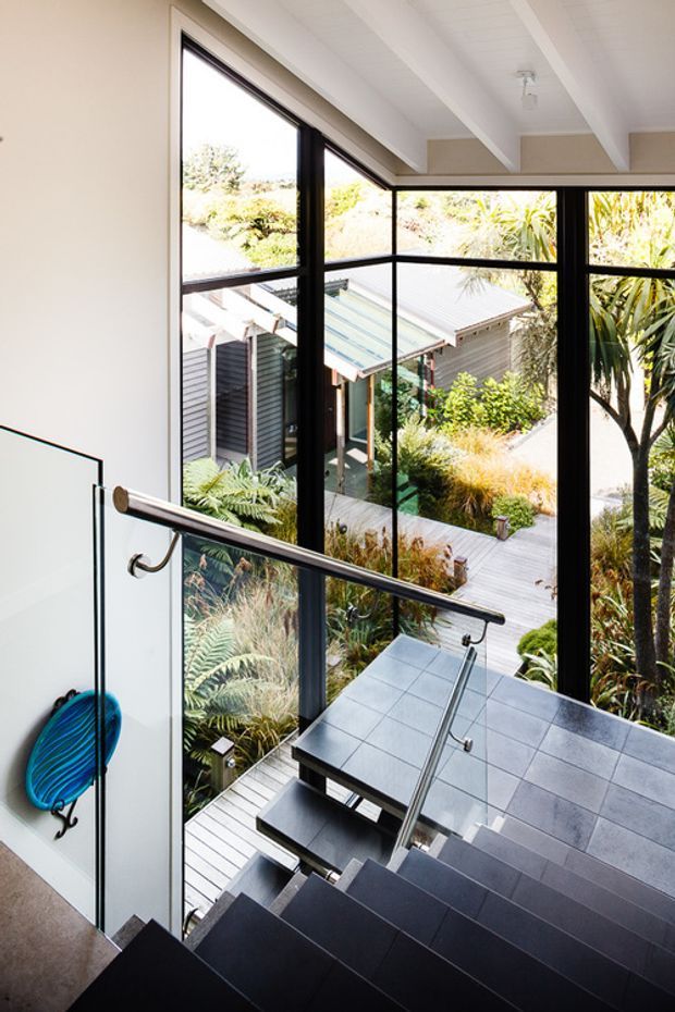 A bright contemporary stairway with dark tiled or timber treads, a glass balustrade, and a polished metal handrail. Floor-to-ceiling black-framed glazing overlooks a landscaped courtyard with native planting and a connected garden walkway. The white pitched ceiling and strong indoor-outdoor connection create an airy, modern atmosphere.