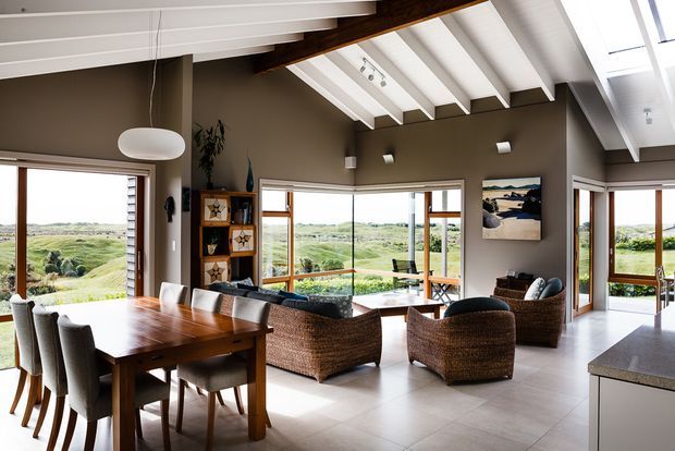 A bright, open-plan dining and living area with a timber dining table and upholstered chairs in the foreground, woven lounge chairs and sofa beyond, and expansive windows overlooking a rural landscape. The room features a dramatic pitched ceiling with exposed dark timber beams, white rafters, skylights, warm taupe walls, pale tiled flooring, and timber-framed glazing. A partial kitchen island is visible at right.