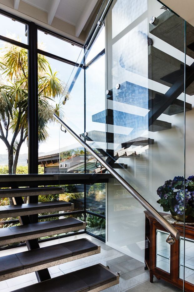 A bright contemporary stairway with open-riser floating timber or dark-finished steps, a polished metal handrail, and frameless glass balustrades. Full-height glazing brings in abundant daylight and overlooks lush planting and a residential setting. A white structural wall and exposed stair support create a clean, airy architectural composition.