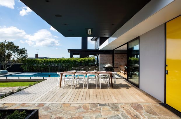 A contemporary outdoor entertaining terrace beside a swimming pool, with a long timber dining table and chairs, large glazed openings, a bright yellow entry door, and a broad dark roof overhang. Natural stone paving borders the timber deck, while hedges and blue sky surround the modern home.