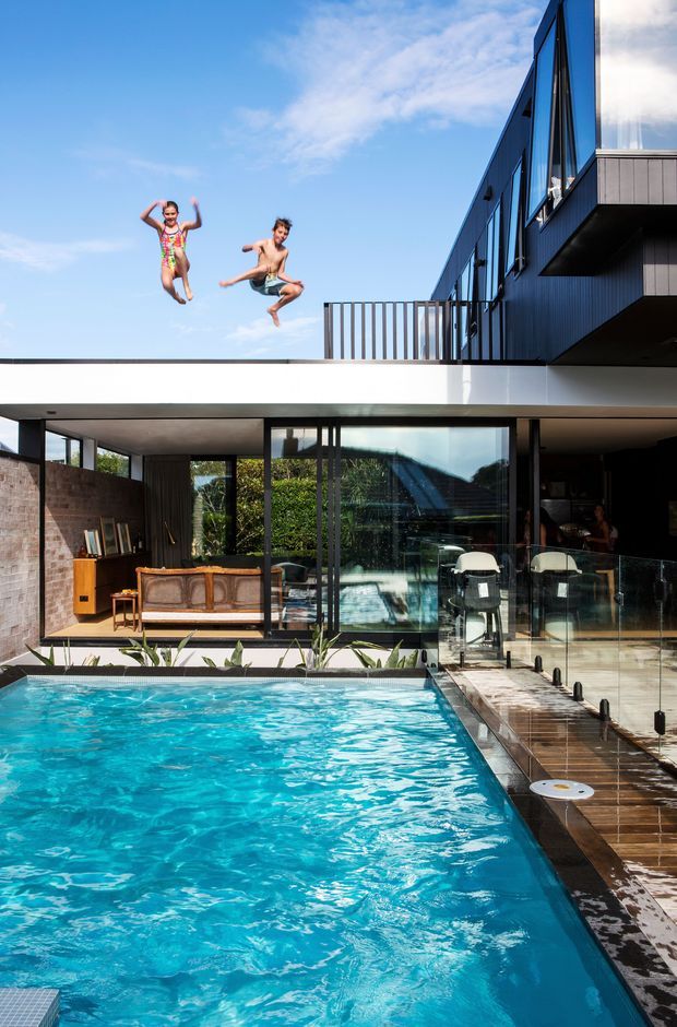A contemporary multi-level house centered around a large turquoise swimming pool. The image shows a dark-clad upper storey with extensive glazing and projecting volumes, a white flat-roofed lower structure, brick feature walls, a glass balustraded pool deck, and open-plan living areas visible behind the pool. Two people are captured jumping above the roofline, emphasizing the recreational setting.