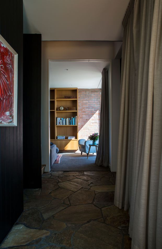 A dim, atmospheric interior hallway or entry passage leading into a small sitting room. The foreground has irregular natural stone flagstone flooring, dark vertical timber wall panelling on the left, and full-height taupe curtains on the right. Beyond the doorway, a warm timber built-in bookshelf, exposed red brick wall, blue upholstered armchair, small round side table, and soft rug create a cozy reading or lounge area. Natural sunlight falls across the brick wall and furniture, creating strong light-and-shadow contrast.