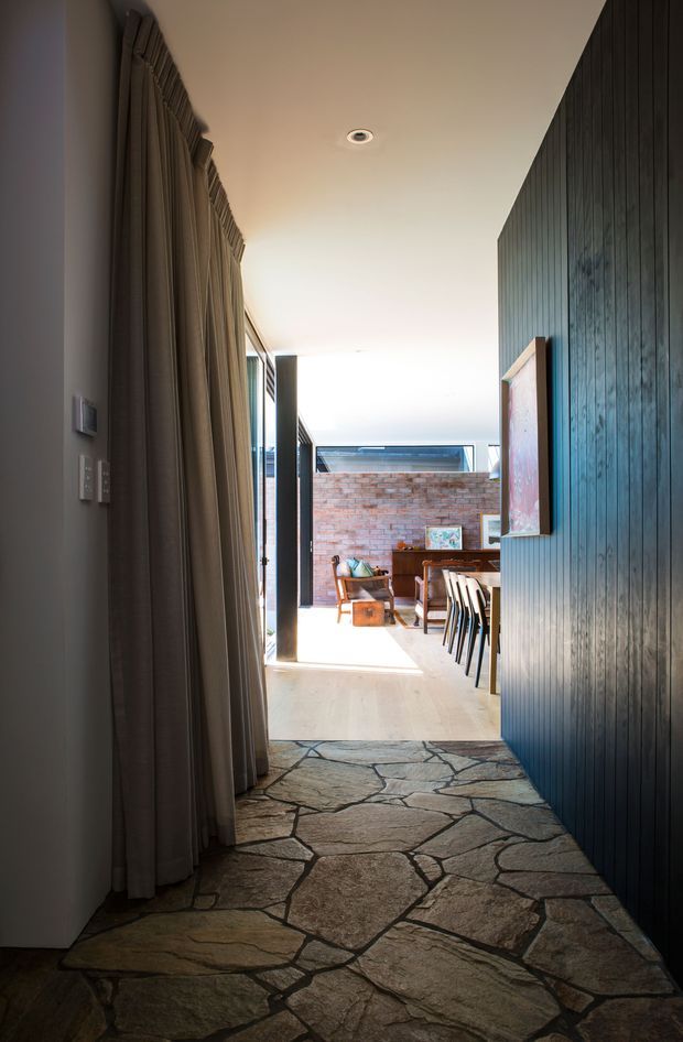 A narrow, dimly lit interior hallway leads into a bright dining and living area. The corridor has an irregular natural stone floor, a dark vertical timber-panelled wall on the right, full-height taupe curtains on the left, and a smooth light ceiling with recessed lighting. Beyond the hallway is a sunlit open space with exposed brickwork, timber furniture, dining chairs, artwork, and glazing or an open connection to an outdoor courtyard.