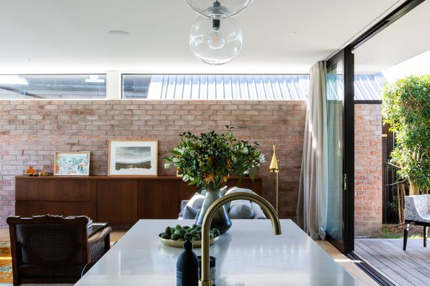 Bright contemporary kitchen-dining space centred on a large white island with an integrated curved brass-toned tap and potted greenery. A warm timber sideboard, exposed brick wall, framed artwork, pendant lighting, and full-height black-framed glazing opening to a timber deck create a refined indoor-outdoor connection.