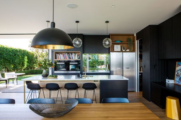 A contemporary open-plan kitchen and dining area with a large central island, four bar stools, dark built-in cabinetry, stainless-steel refrigerator, timber shelving, and a light stone benchtop. Black dome pendant lights hang above the island, while full-height glazing opens to a leafy outdoor area. A timber dining table occupies the foreground.