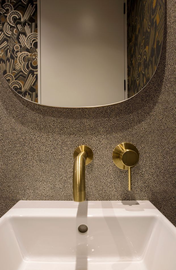 Close-up view of a refined bathroom vanity featuring a white rectangular basin, wall-mounted brushed brass cylindrical tapware, and a matching brass control. The wall is covered in a dark, highly patterned textured finish resembling small circular beads. A large curved-edge mirror reflects bold geometric Art Deco-style wallpaper and dark patterned surfaces, creating a luxurious, dramatic composition.