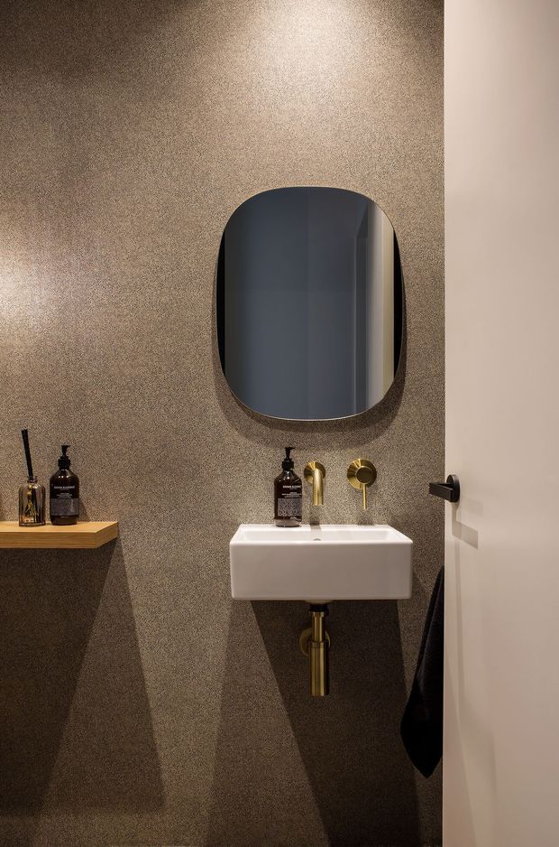 Compact, refined bathroom featuring a wall-mounted white rectangular basin with exposed brass-gold plumbing, matching brass-gold wall-mounted tapware, and a large rounded-rectangle dark mirror. The walls and floor are finished in a textured dark taupe-brown surface, with a floating timber shelf and toiletries at left. A white door with dark hardware is visible at right. Warm directional lighting creates a moody, boutique-hotel atmosphere.
