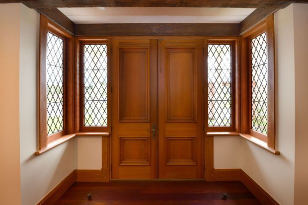 A formal residential entryway with a richly stained timber double front door, matching panelled timber surround, and narrow diamond-leaded glass sidelights. The space has white plaster walls, dark timber flooring and skirting, and exposed rustic ceiling beams, creating a traditional, warm, heritage character.