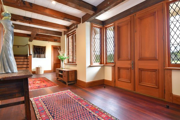 Traditional residential entry hall featuring richly stained timber panelled double doors, matching diamond-leaded windows, exposed dark timber ceiling beams, polished timber flooring, warm cream walls, and Persian-style runners. A staircase, console cabinet, artwork, and decorative sculpture reinforce the formal heritage character.