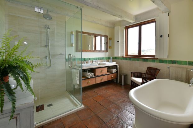 Spacious traditional bathroom with a glass-enclosed walk-in shower, freestanding curved bathtub, double vanity, wood-framed windows, and terracotta-toned floor tiles. Green accent tiles form a horizontal border around the room, while exposed beams and timber cabinetry add warmth.