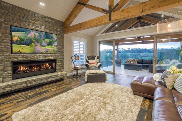 Spacious rustic-contemporary living room with exposed timber beams, a vaulted white-paneled ceiling, and a stacked-stone feature wall containing a long linear fireplace and television. Leather sectional seating, armchairs, a large textured rug, and warm timber flooring create a comfortable lounge setting. Full-height glazed sliding doors open to a covered outdoor area and scenic landscape beyond.