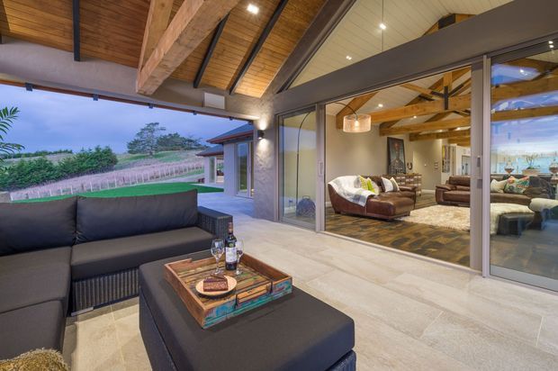 A spacious covered outdoor lounge and adjoining living room are connected by wide sliding glass doors. The outdoor area has a dark sectional sofa and ottoman around a rustic timber coffee table, while the interior features leather seating, timber beams, a pitched timber-lined ceiling, and a large rug. The setting opens toward landscaped grounds and vineyards, creating a relaxed indoor-outdoor entertaining space.