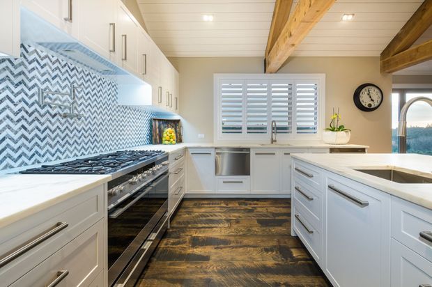 Bright, spacious kitchen with white shaker-style cabinetry, a large central island, stainless-steel range and appliances, a bold blue-and-white chevron tile splashback, white stone benchtops, dark timber flooring, and exposed timber ceiling beams. Plantation shutters and a view through the window add a relaxed residential feel.