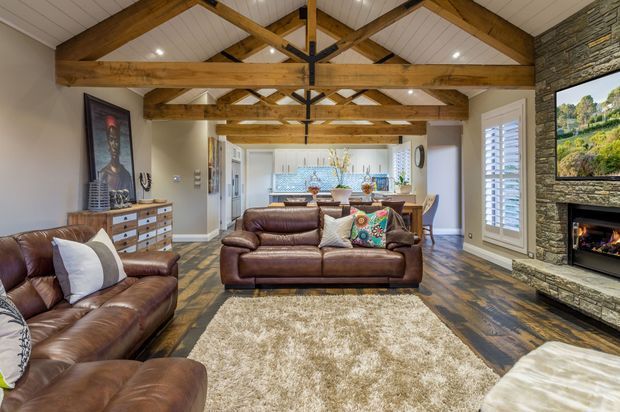 A warm, spacious open-plan living room with exposed timber trusses and beams, leather sofas, a textured area rug, and a stone-clad fireplace wall with television. The room opens toward a bright kitchen and dining area, with white plantation shutters and polished timber flooring.