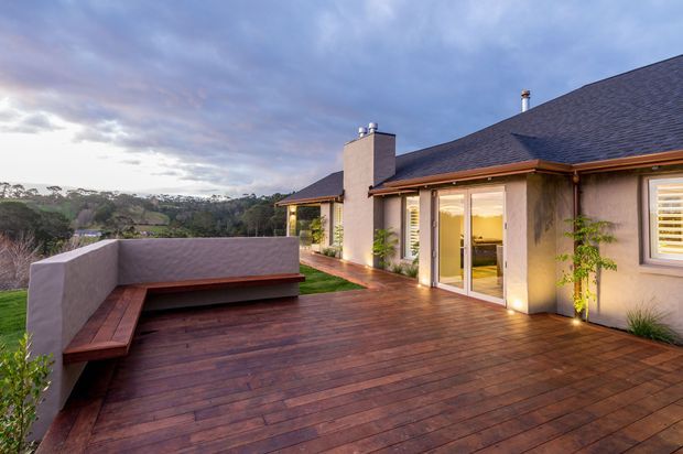A spacious elevated timber deck beside a single-storey home, featuring built-in bench seating along a rendered privacy wall, warm exterior lighting, large glazed sliding doors, and a pitched dark shingle roof. The deck overlooks a leafy rolling landscape at dusk.