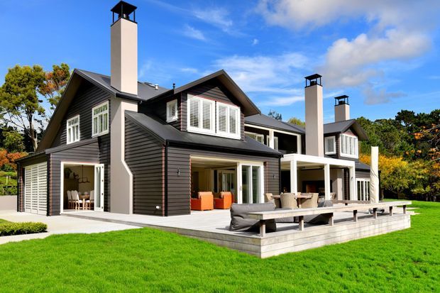 A large multi-level suburban residence with dark horizontal cladding, multiple steep gabled roof sections, tall pale chimneys with black caps, white plantation shutters, and broad glazed openings. A generous paved terrace with outdoor dining and lounge furniture overlooks a manicured lawn, while mature trees surround the property.