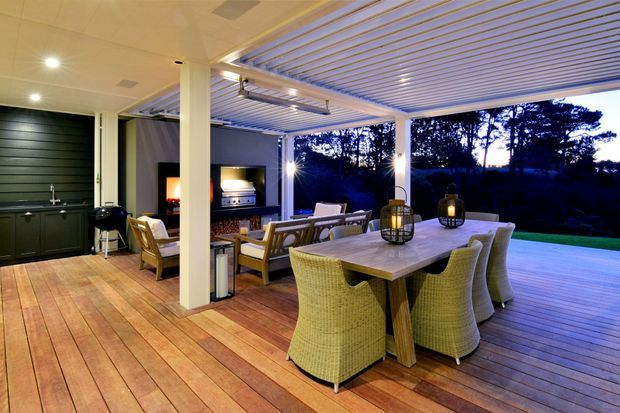 A covered outdoor entertaining area with a timber deck, dining table and woven dining chairs, lounge seating, built-in outdoor kitchen and barbecue, and a louvred pergola roof. The space opens onto a landscaped lawn and wooded outlook at dusk, with integrated lighting creating a warm, inviting atmosphere.