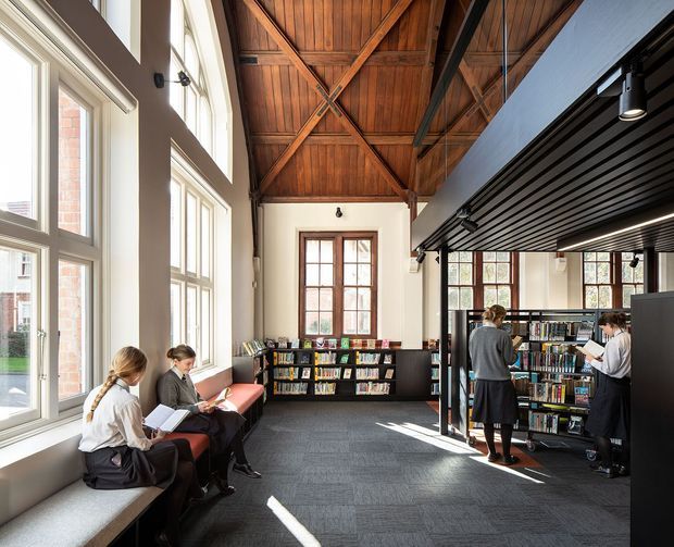 Bright double-height library interior with tall multi-pane windows, built-in bookshelves, students reading and browsing books, and a dramatic exposed timber truss ceiling. The space combines a historic-looking masonry shell with a dark contemporary slatted mezzanine or upper-level structure and track lighting.
