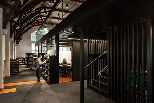 Interior view of a contemporary library or reading facility, combining a double-height open reading area with bookshelves, study seating, and a dramatic black metal stair enclosure. Exposed dark timber roof trusses, clerestory glazing, linear ceiling lighting, and charcoal vertical metal battens create a strong industrial and adaptive-reuse character. The foreground is dominated by a stairway and circulation zone, while the library space is visible beyond.